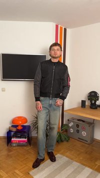 Black bomber jacket with gray sleeves, zipper closure, and sleeve patch. Light blue straight-leg jeans and brown leather shoes. Indoor setting.