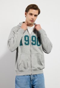 Grå zip-up hettegenser med frontlomme og stor grønn "1990" print. Myk stoff med hette og ribbestrikkede mansjetter.