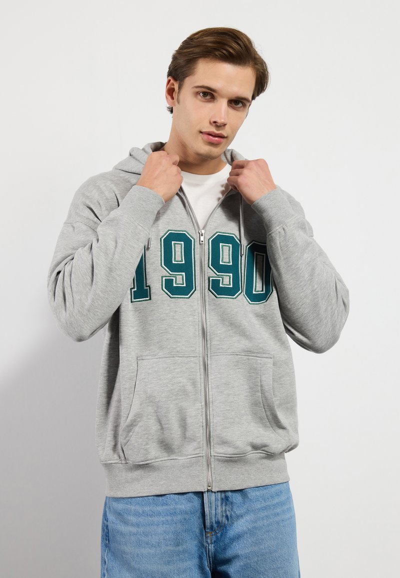 Grå zip-up hettegenser med frontlomme og stor grønn "1990" print. Myk stoff med hette og ribbestrikkede mansjetter.