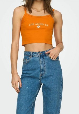 Mladá žena v oranžovém krátkém topu s nápisem "LOS ANGELES" a vysokým pasem modrých džínů, s jednou rukou v kapse, na bílém pozadí.