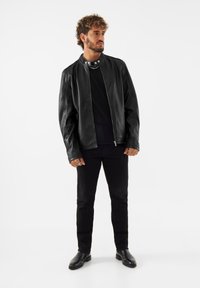 Veste en cuir noir avec un col montant, fermeture éclair et manches longues. Portée par-dessus une chemise noire et assortie à un pantalon noir et des bottes.