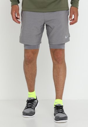 Mann trägt graue Lagen-Shorts, neongelbe Nike-Socken, schwarz-weiße Laufschuhe und steht auf einem weißen Boden.