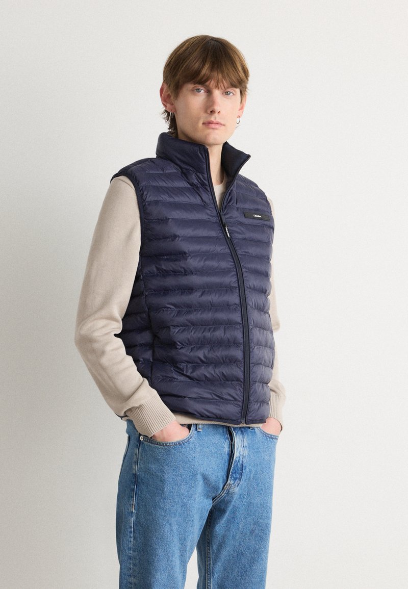 Jūras spēku pūkains vestes ar augstu apkakli, ar rāvējslēdzēju un horizontālu quilted rakstu. Valkāta virs bežīgas džemperes un ziliem džinsiem.