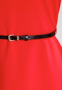 Cinturón delgado negro con hebilla dorada llevado alrededor de la cintura sobre un vestido o prenda de color rojo brillante.