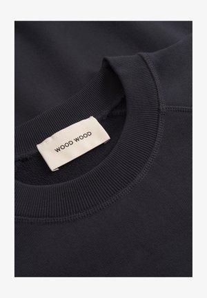 Približan posnetek ovratnika temno modre rebraste pulover majice z okroglim izrezom, na notranji strani izreza je prišita bela etiketa blagovne znamke "WOOD WOOD".