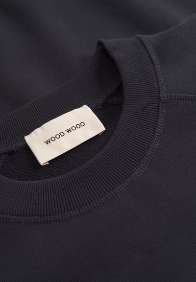 Približan posnetek ovratnika temno modre rebraste pulover majice z okroglim izrezom, na notranji strani izreza je prišita bela etiketa blagovne znamke "WOOD WOOD".