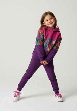 Fioletowy dres z różową bluzą z kapturem ozdobioną wzorem roślinnym. Białe trampki z różowymi sznurowadłami. Gładka faktura tkaniny.