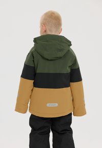 Kleurgebloemde jas met een groen, zwart en tan ontwerp. Voorzien van een capuchon en reflecterende accenten. Gedragen over zwarte broek.