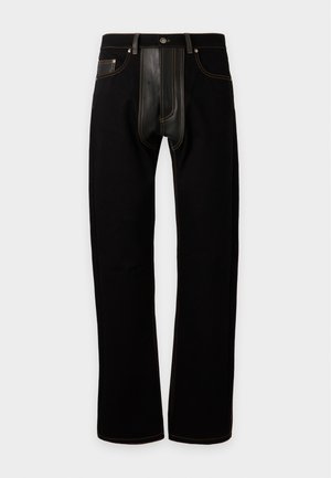 Mann trägt ein buntes Langarmhemd mit abstrakten Mustern und schwarze Hose mit schwarzen Loafern mit goldenen Spitzen, steht vor weißem Hintergrund.