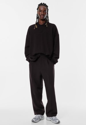 Oversized zwarte polo met een kraag, gecombineerd met losse zwarte broek. Grijze sneakers met een gestructureerd patroon maken de outfit af.