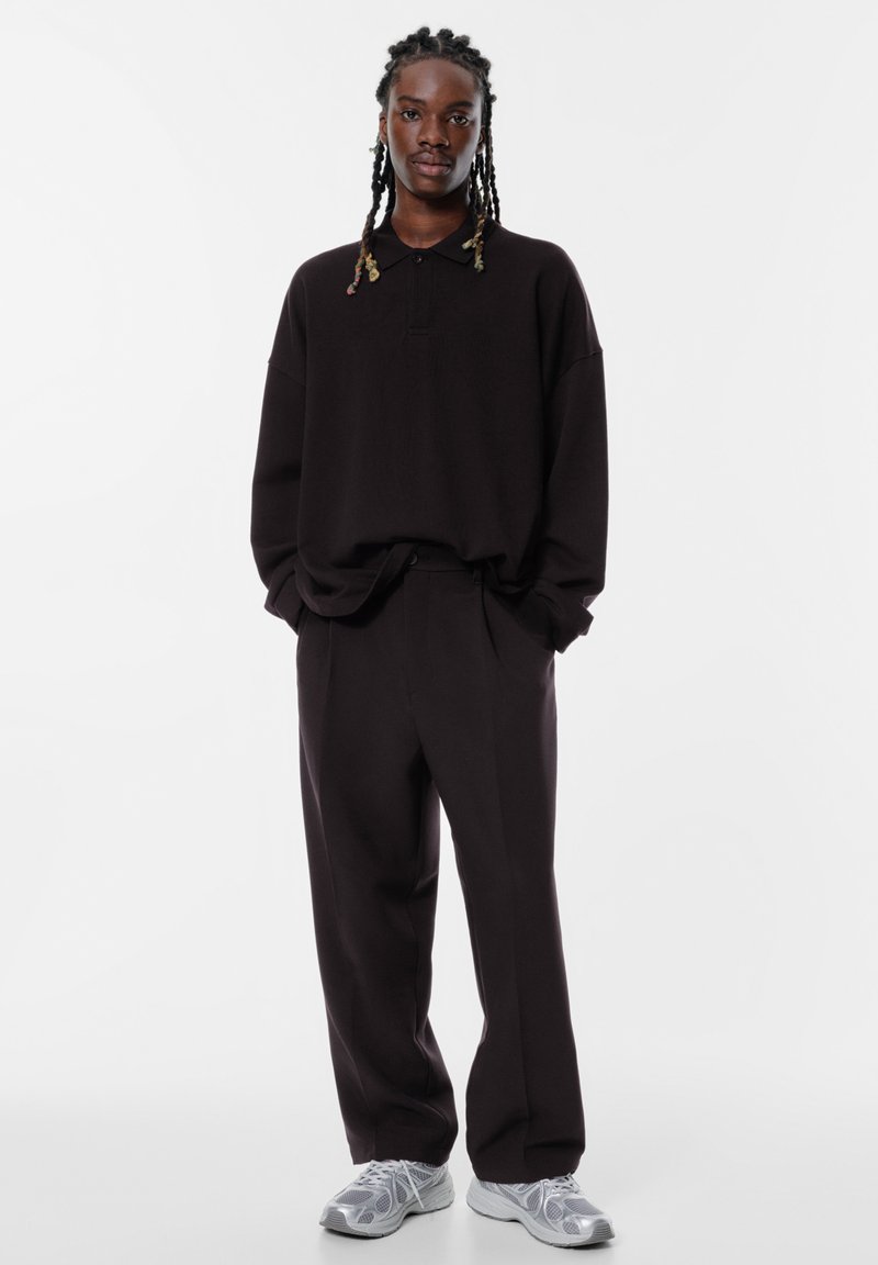 Überdimensioniertes schwarzes Poloshirt mit Kragen, kombiniert mit lockeren schwarzen Hosen. Graue Sneaker mit strukturiertem Muster vervollständigen das Outfit.
