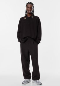 Oversized zwarte polo met een kraag, gecombineerd met losse zwarte broek. Grijze sneakers met een gestructureerd patroon maken de outfit af.