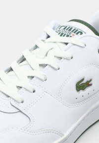 Fehér bőr sneaker zöld kiegészítőkkel. Hímzett logóval, struktúrált fűzőkkel és perforációkkal a lábujjak közelében a légáteresztés érdekében.