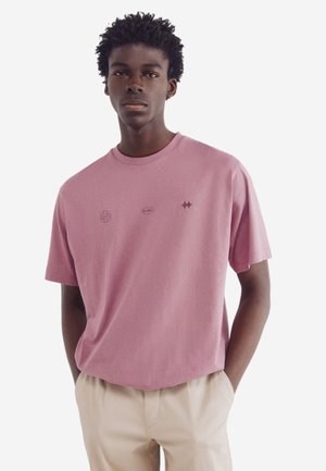 Lyserød bomulds t-shirt med korte ærmer, rund hals, der har tre små grafiske elementer i mørkeblå og sort på brystet.