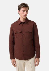 Braune, knöpfbare Jacke mit langen Ärmeln, zwei Brusttaschen und einem spitzen Kragen. Glatte Textur und minimalistische Designakzente.