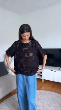 Femme à l’intérieur ajustant un haut noir transparent à motifs floraux avec un ourlet volanté, portant un jean bleu ample, debout sur un tapis rond clair près d’une télévision et d’un canapé.