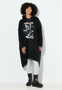 Zwarte capuchon-tuniek met een grafische print van een kat en muziek elementen. Gemaakt van zachte stof, met een langere achterkant en zilveren accenten.