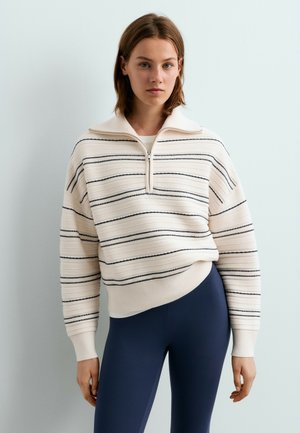 Jonge vrouw draagt een witte trui met rits met zwarte horizontale strepen en donkerblauwe leggings tegen een effen lichte achtergrond.