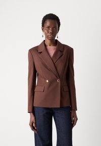 Dobbeltspent brun blazer i strukturert stoff, med spiss krage, to gullknapper og frontlommer, iført over en rosa topp.