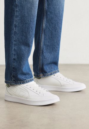 White sneakers with lace-up design, perforated accents, and a smooth texture, paired with blue denim jeans, showcasing a relaxed fit.