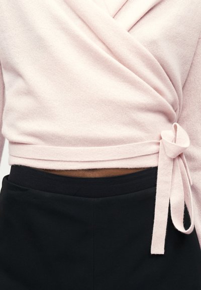 Jasnoróżowy sweter wiązany w talii, zestawiony z czarnymi spodniami z wysokim stanem, odsłaniając niewielki fragment brzucha.