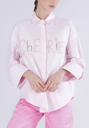 Donna che indossa una camicia rosa chiaro con bottoni, con la scritta "CHERIE" realizzata con decorazioni di perle, abbinata a pantaloni rosa coordinati.