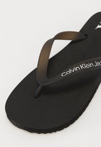 Fekete papucs szövet talppal és rugalmas sötétbarna pánttal. A pánton fehér "Calvin Klein Jeans" márkázás található.