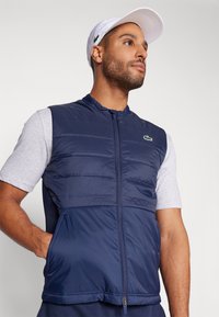 Colete acolchoado azul-marinho com fecho éclair, apresentando um detalhe de logotipo verde. Usado sobre uma camisa de manga curta cinza, com bolsos laterais. Textura de tecido suave.