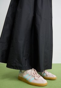 Beige och ljusblå sneakers med gummisulor, som bärs med vita strumpor, delvis täckta av en lång svart kjol på ett grönt golv.