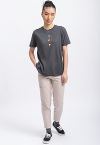Grijze T-shirt met korte mouwen en drie kleine versieringen, gecombineerd met lichtbeige broeken en zwarte sneakers. Model staat met handen in de zakken.
