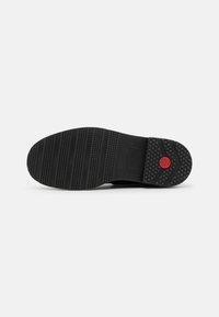 Semelle de chaussure en caoutchouc noir avec des grips texturés, présentant des rainures horizontales et verticales, et un logo circulaire rouge au niveau du talon.