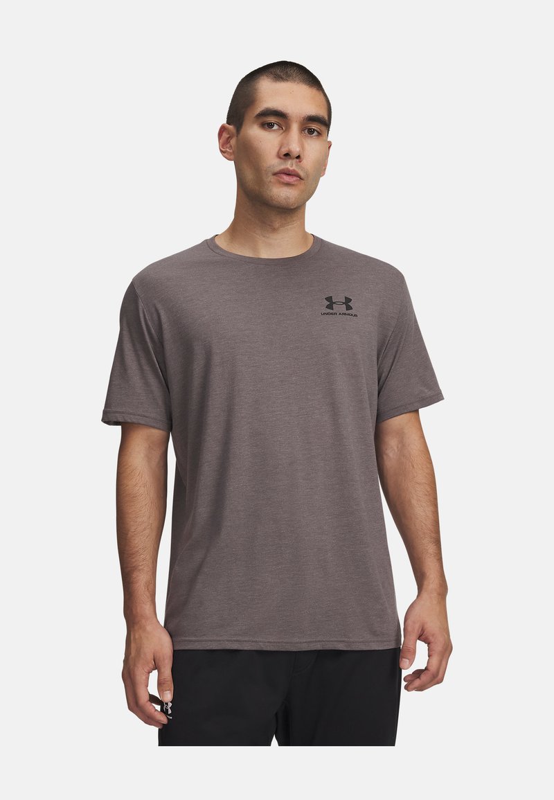 Мъж, облечен с обикновена сива тениска с къси ръкави с черно лого на Under Armour на гърдите и черни къси панталони на бял фон.