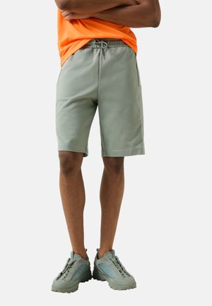 Ein Mann steht mit verschränkten Armen und trägt hellgrüne Shorts mit Kordelzug, ein oranges Shirt und passende hellgrüne Turnschuhe.