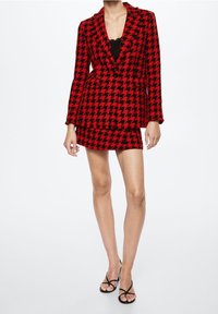 Rood en zwart houndstooth-patroon blazer met een cinched taille, gelaagd over een zwarte top, gecombineerd met een bijpassende mini-rok en zwarte hakken.