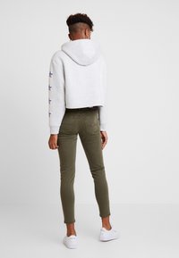 Persona che indossa un hoodie corto grigio chiaro con disegni a stella sulle maniche, jeans skinny verde oliva e sneakers bianche, in piedi girato di spalle.