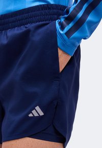 Personne portant un short de sport bleu foncé avec un logo blanc et une main dans la poche, associé à un haut bleu à manches longues avec des rayures.