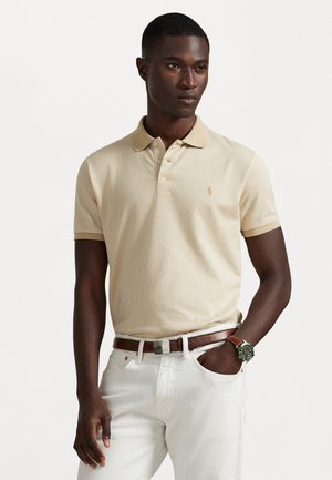 Beige polo shirt met een gestructureerd patroon, korte mouwen en een kraag; heeft een klein gouden logo op de linkerborst; gecombineerd met witte broeken.