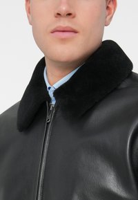 Fekete bőrkabát fekete gyapjú gallérral, elöl cipzárral, egy világoskék ing fölött viselve. Simább textúra.