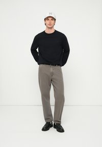 Μαύρο πουκάμισο με μακριά μανίκια, γκρι παντελόνι και μαύρα παπούτσια. Το άτομο φοράει λευκό καπέλο με τετράγωνο λογότυπο στο μπροστινό μέρος.