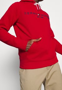 Homme portant un hoodie rouge Tommy Hilfiger avec des cordons blancs et un pantalon beige, la main dans la poche du hoodie, sur un fond uni.