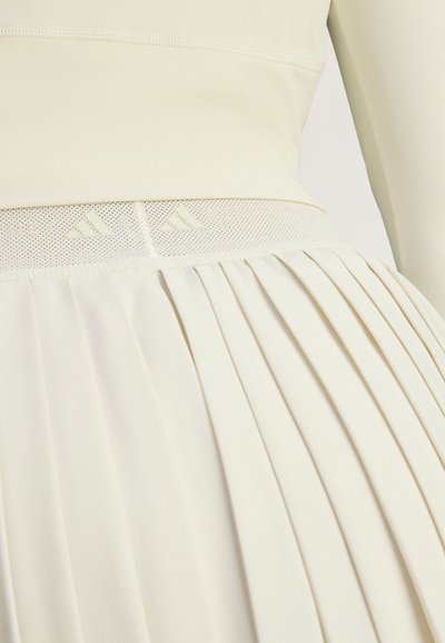 Light cream dress featuring a fitted top with a mesh panel at the waist, and a pleated skirt with a smooth texture.