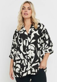 Blusa bianca e nera con motivi geometrici astratti, maniche lunghe con polsini arricciati e chiusura con bottoni. Tessuto leggero.