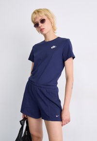 T-shirt e calções em mistura de algodão, de cor azul-marinho da Nike, com um logo branco no peito. Acessórios incluem óculos de sol redondos e uma mala preta.
