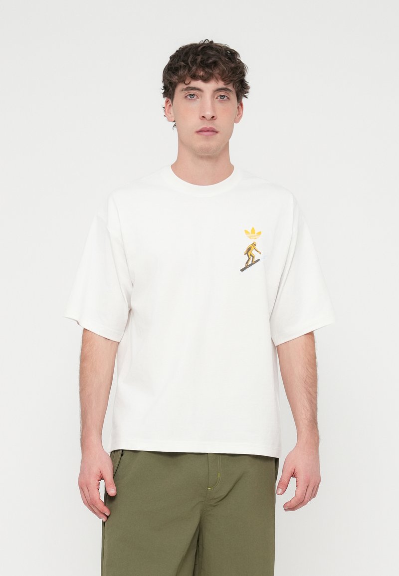 Vit bomulls-T-shirt med korta ärmar, med ett tryck av en gul karaktär och logotyp på vänster bröst. Avslappnad passform, slät tygstruktur.