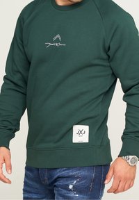 Uomo che indossa una felpa Jack & Jones verde scuro con logo sul petto e sull'orlo, abbinata a jeans blu strappati e un orologio da polso in argento.
