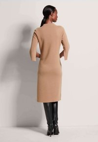 Strickkleid in Camel mit 3/4-Ärmeln, knieumspielend, glatte Textur. Kombiniert mit hohen, schwarzen, körperbetonten Stiefeln mit spitzem Zehen.