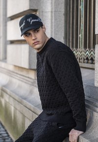 Suéter negro de punto con un patrón texturizado y puños acanalados, combinado con pantalones a juego. Una gorra negra presenta texto en cursiva blanca.
