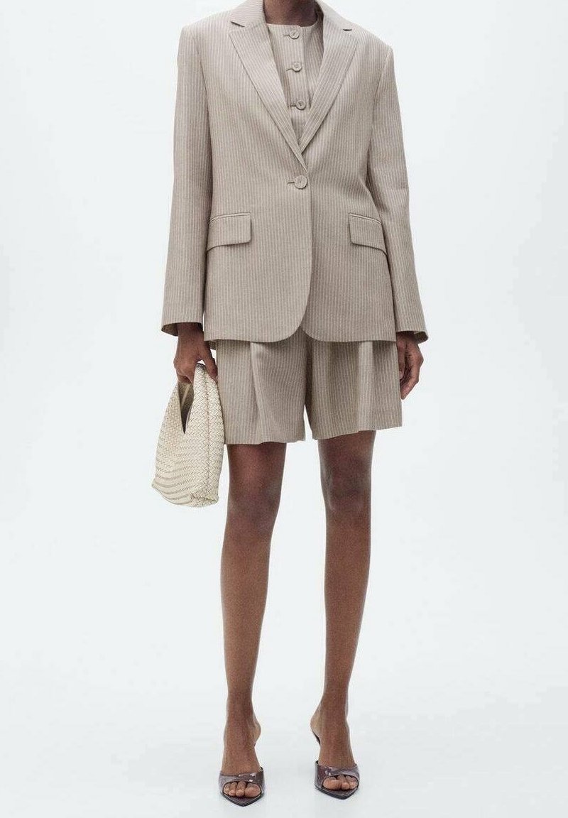 Femme portant un blazer beige à fines rayures, un short assorti, des sandales à talons couleur nude et tenant un sac à main beige texturé.