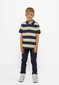 Pruhované polo tričko s krátkým rukávem v béžové a námořnicky modré, s dvěma knoflíky, v kombinaci s námořnickými kalhotami a bílými teniskami.
