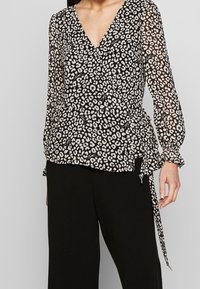 Femme portant un chemisier en imprimé léopard noir et blanc avec de longues manches transparentes, et un pantalon large noir, se tenant devant un fond gris.